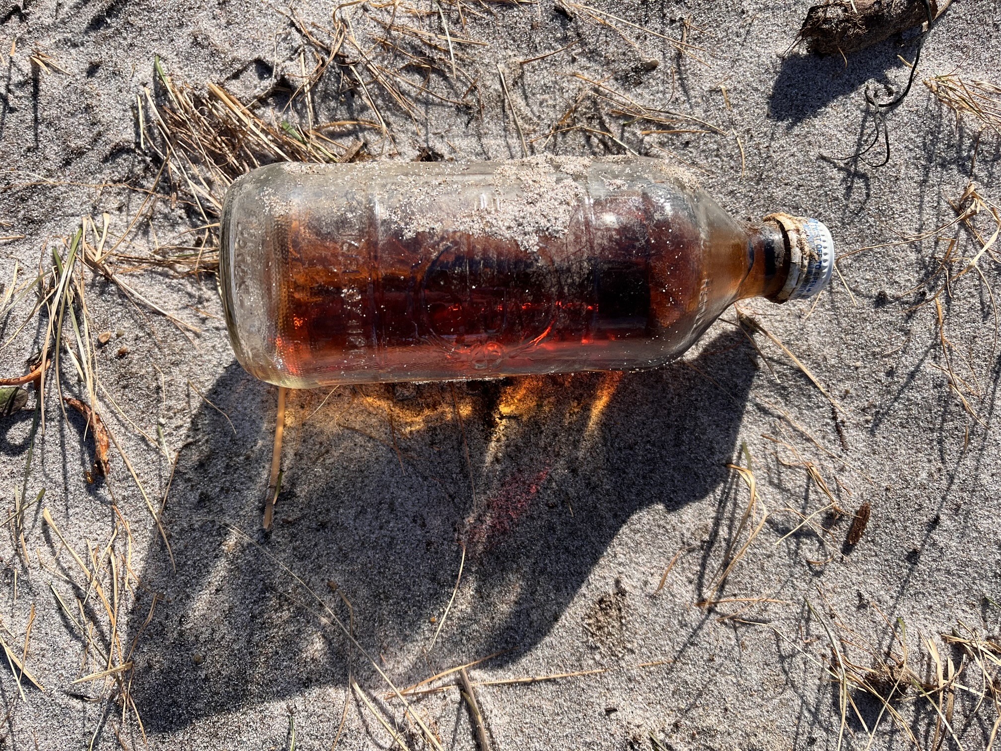An old, clear glass Pepsi bottle, containing some brown liquid, sitting on a sandy beach.